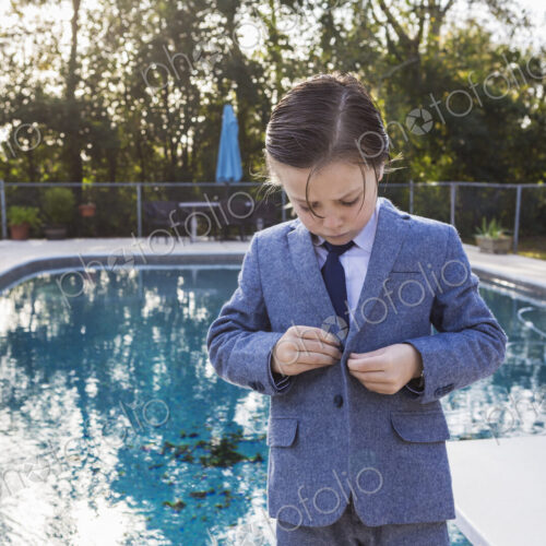6 year old boy buttoning his suit with pool in background 6 year old boy buttoning his suit with pool in background