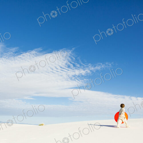 6 year old boy carrying an orange sled in a white undulating dune landscape. 6 year old boy carrying an orange sled in a white undulating dune landscape.