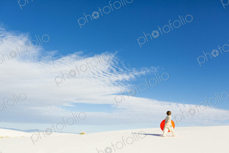 6 year old boy carrying an orange sled in a white undulating dune landscape.