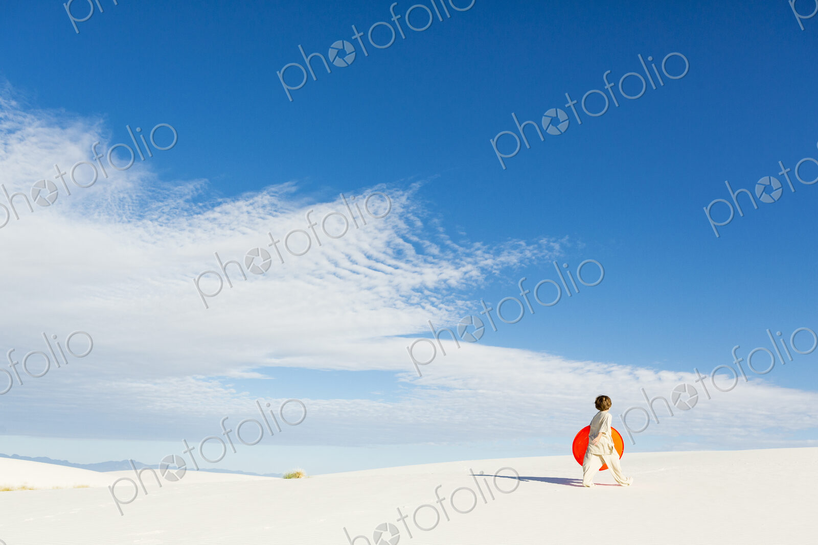 6 year old boy carrying an orange sled in a white undulating dune landscape.