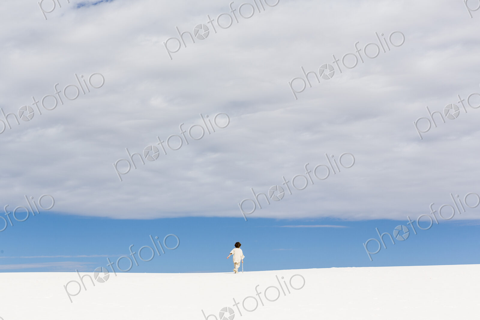 6 year old boy walking in white sand dunes, a strip of blue sky on the horizon and…