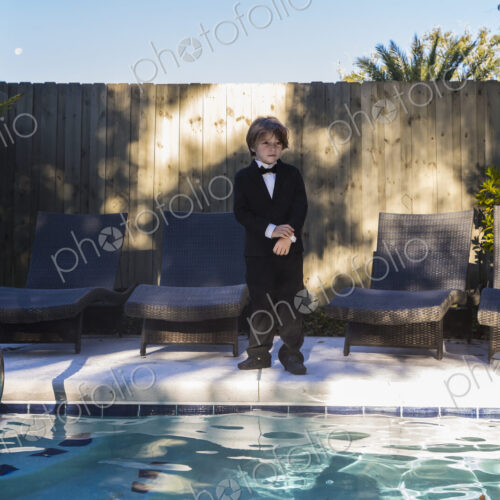 6 year old boy wearing black suit and bow tie standing by a swimming pool