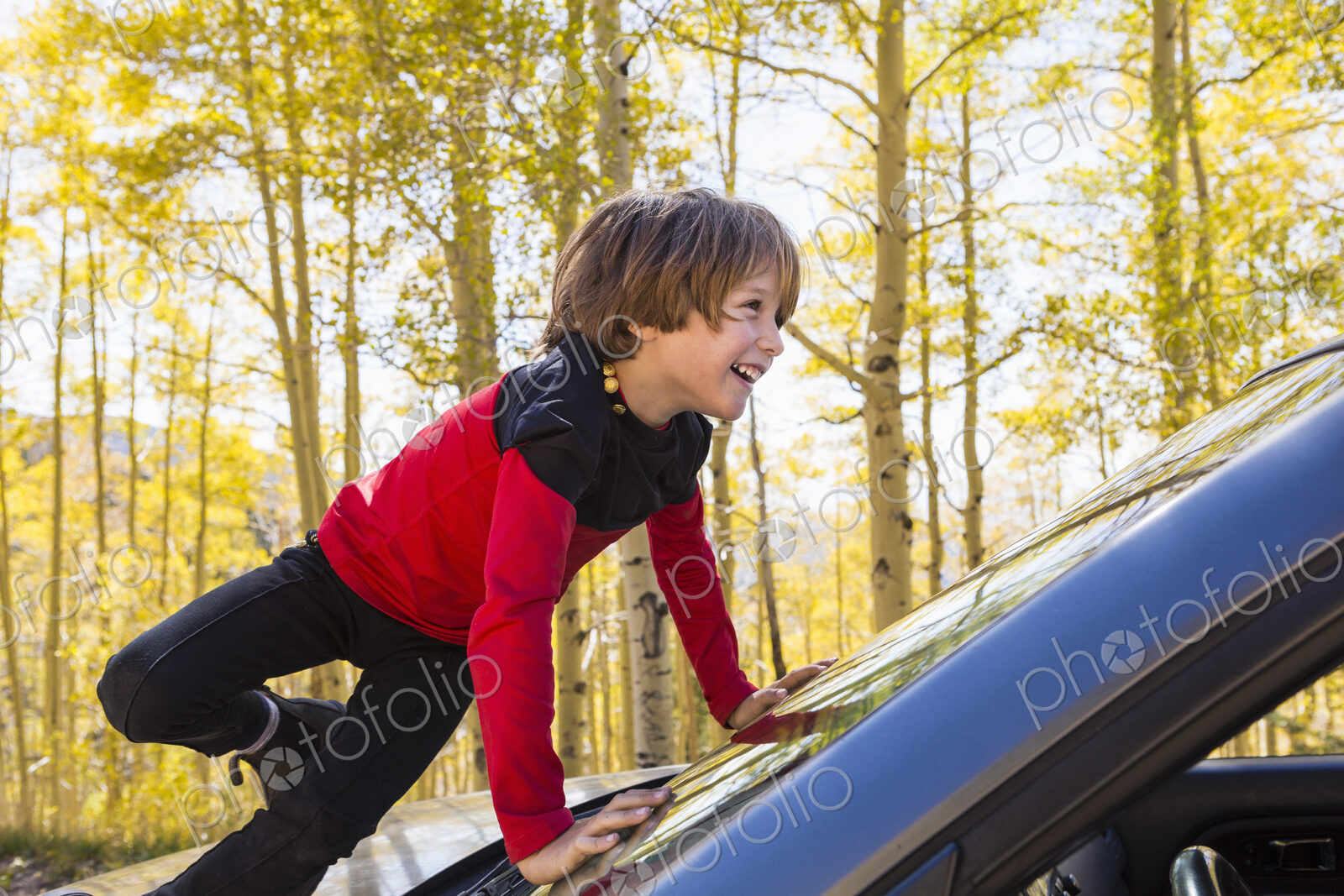 A 6 year old boy climbing on the hood of a blue SUV, among autumn aspens