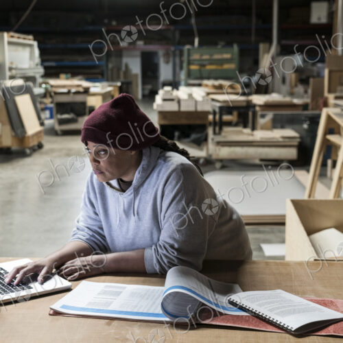 A Black female carpenter working on a lap top computer after work hours in a large… A Black female carpenter working on a lap top computer after work hours in a large…