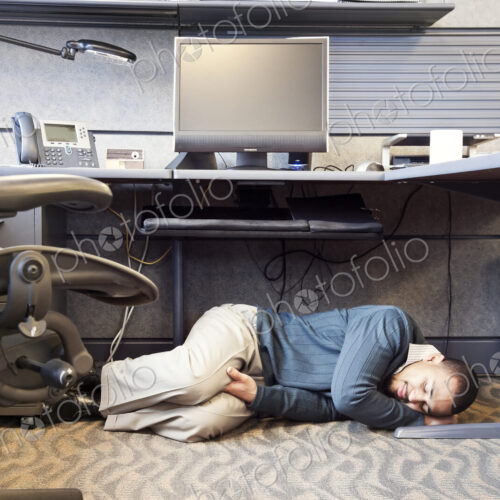 A Black man taking a nap under his desk in a corporate office