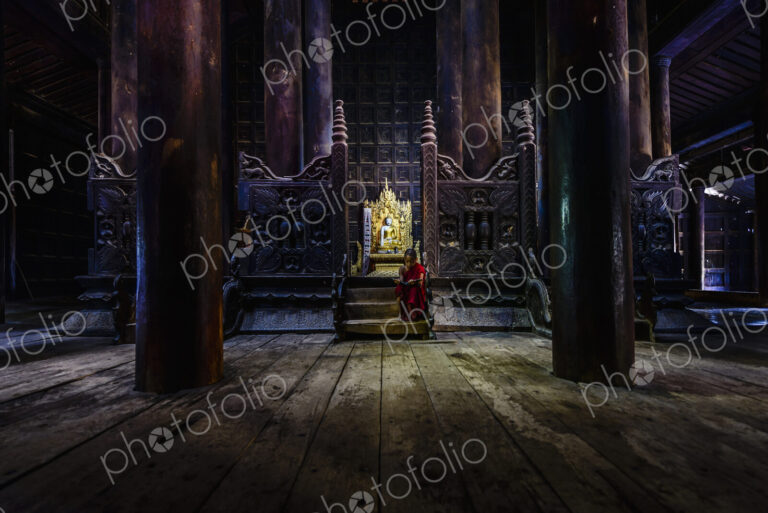 A Buddhist temple with wooden pillars and carved screens, a golden thrine and a young…