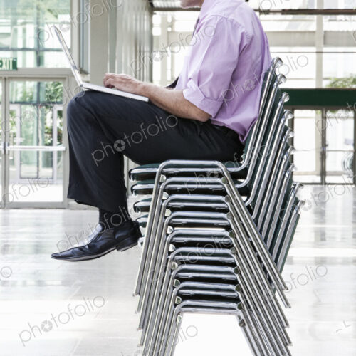 A Caucasian businessman sitting on top of multiple stacked chairs and working on a… A Caucasian businessman sitting on top of multiple stacked chairs and working on a…
