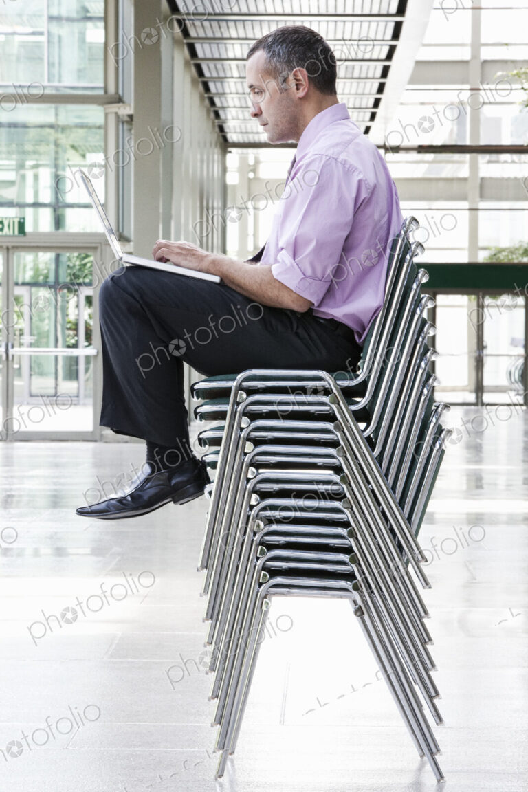 A Caucasian businessman sitting on top of multiple stacked chairs and working on a…