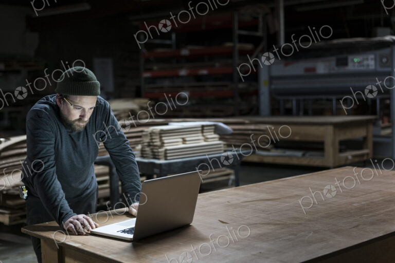 A Caucasian carpenter working on his lap top after hours in a large woodworking factory.