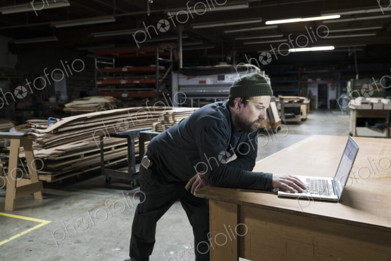 A Caucasian carpenter working on his lap top after hours in a large woodworking factory.