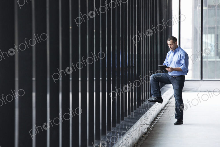 A Caucasian male architect standing by a wall of windows, using a digital tablet.