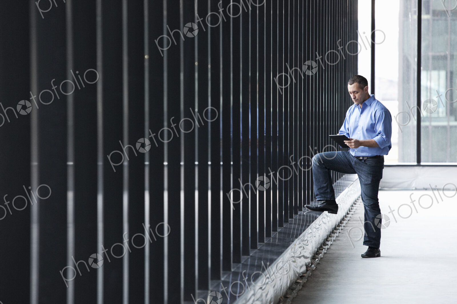 A Caucasian male architect standing by a wall of windows, using a digital tablet.