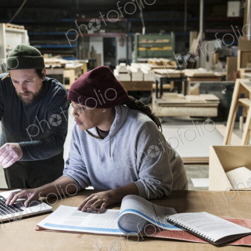 A Caucasian male carpenter and a Black female carpenter working on a laptop computer…