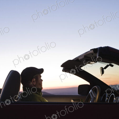 A Caucasian male watching the sunset while parked in his convertible sports car.