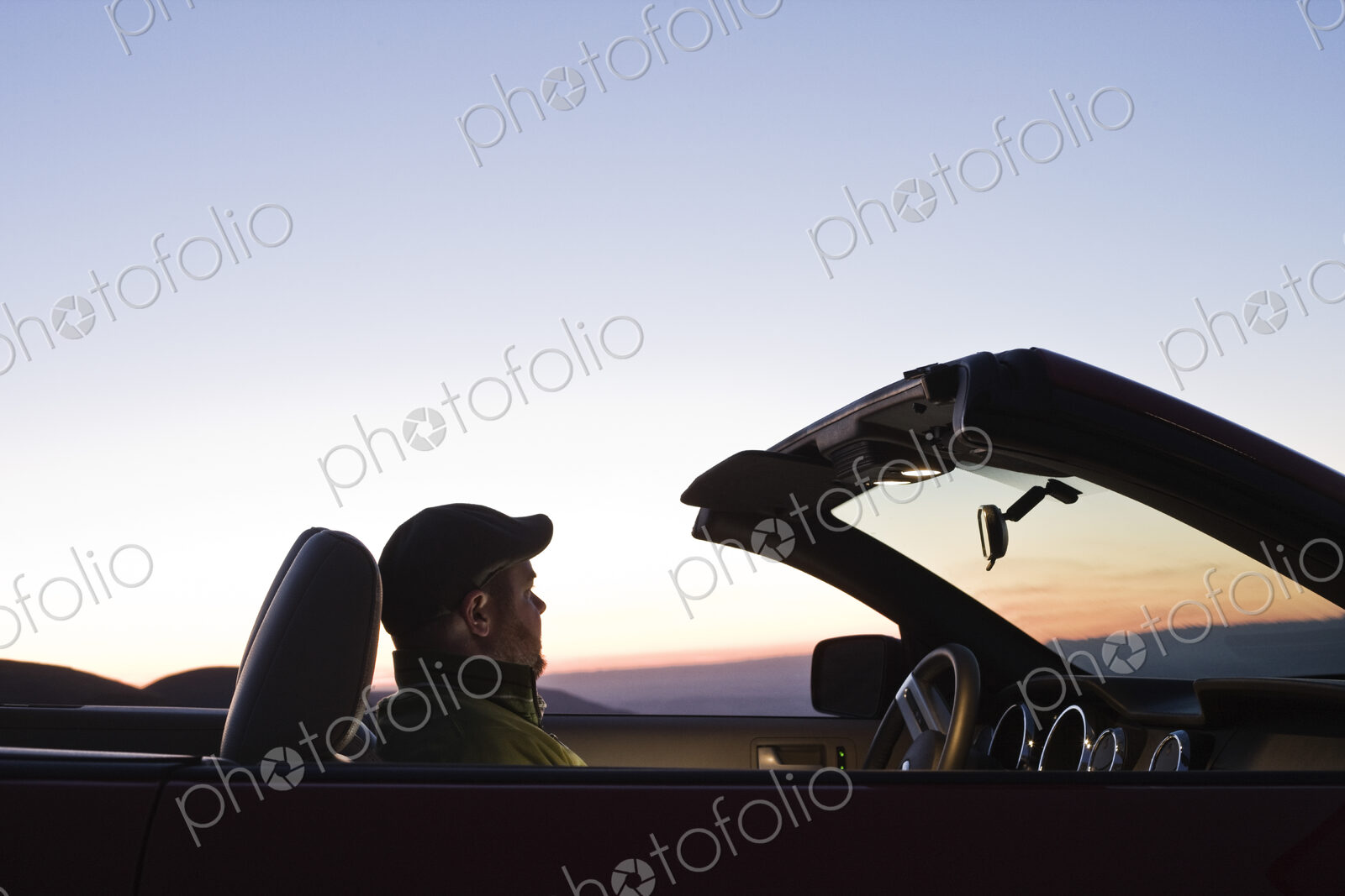 A Caucasian male watching the sunset while parked in his convertible sports car.