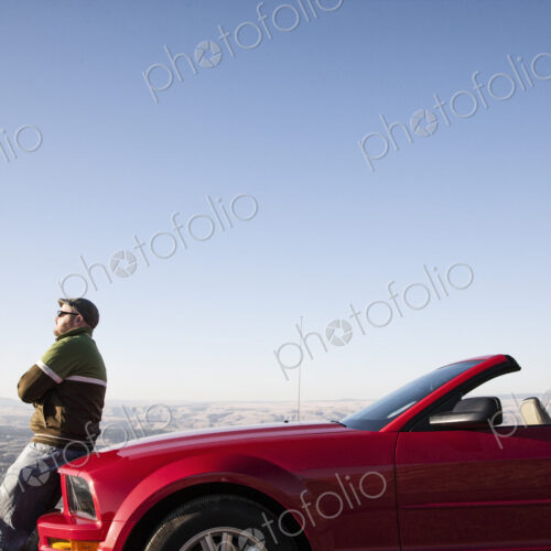 A Caucasian man resting on the hood of his convertible sports car at a rest stop. A Caucasian man resting on the hood of his convertible sports car at a rest stop.