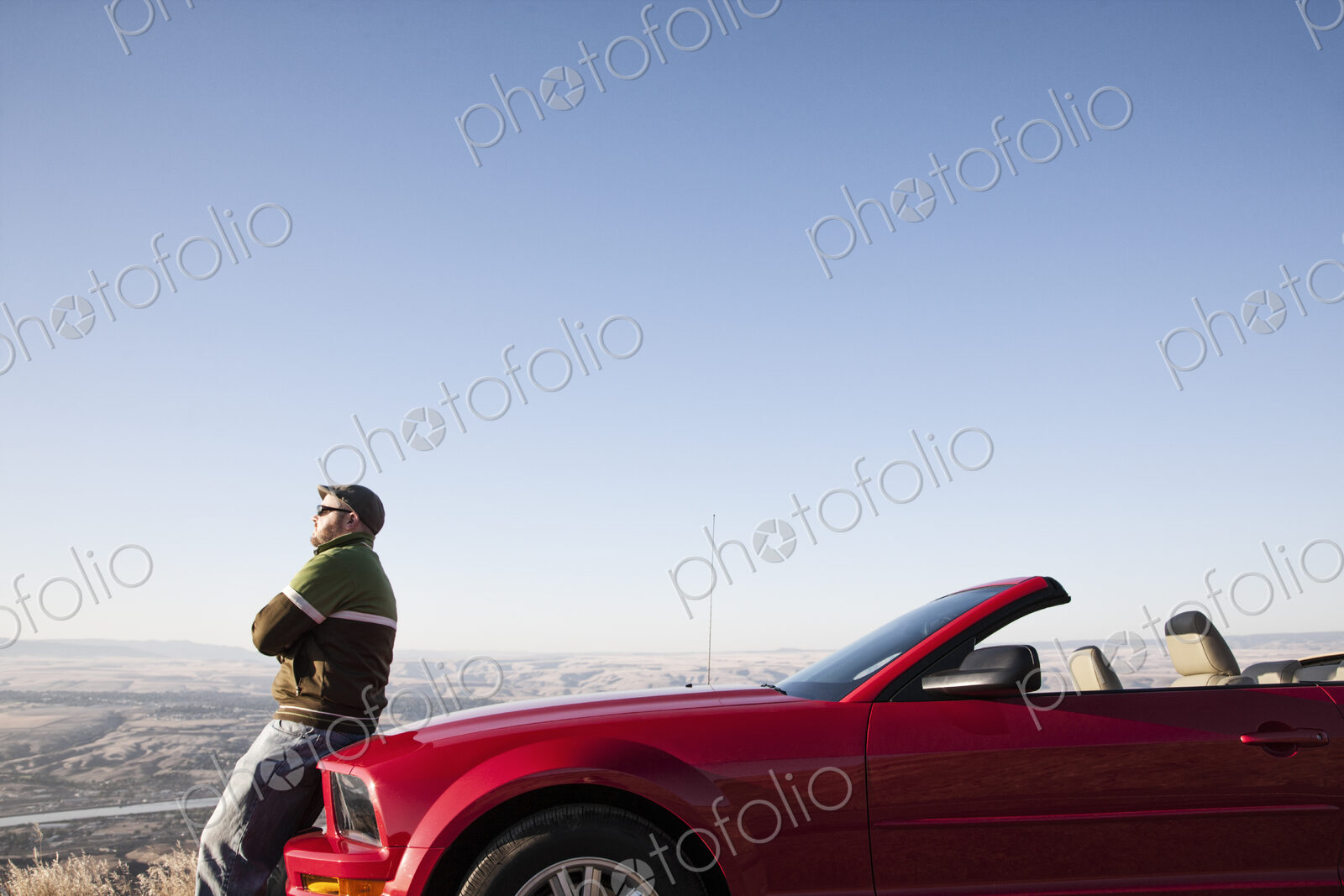 A Caucasian man resting on the hood of his convertible sports car at a rest stop.