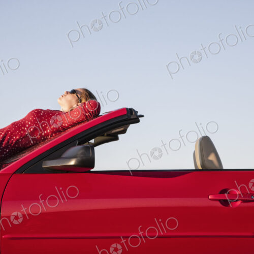 A Caucasian woman resting on the hood of her convertible sports car at a rest stop.
