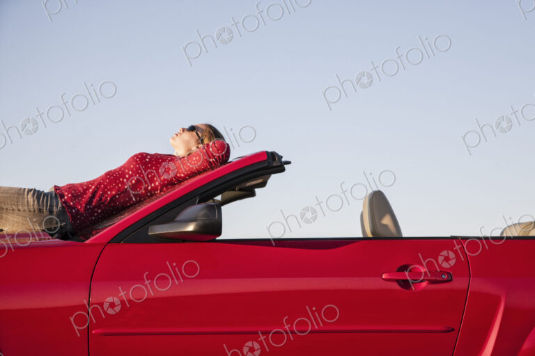 A Caucasian woman resting on the hood of her convertible sports car at a rest stop.