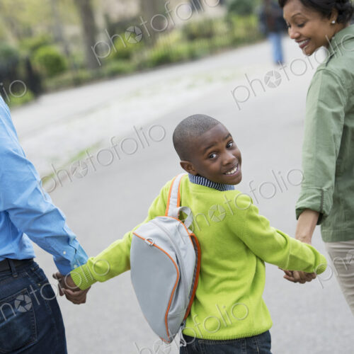 A New York city park in the spring. A boy with a bookbag, holding hands with his…