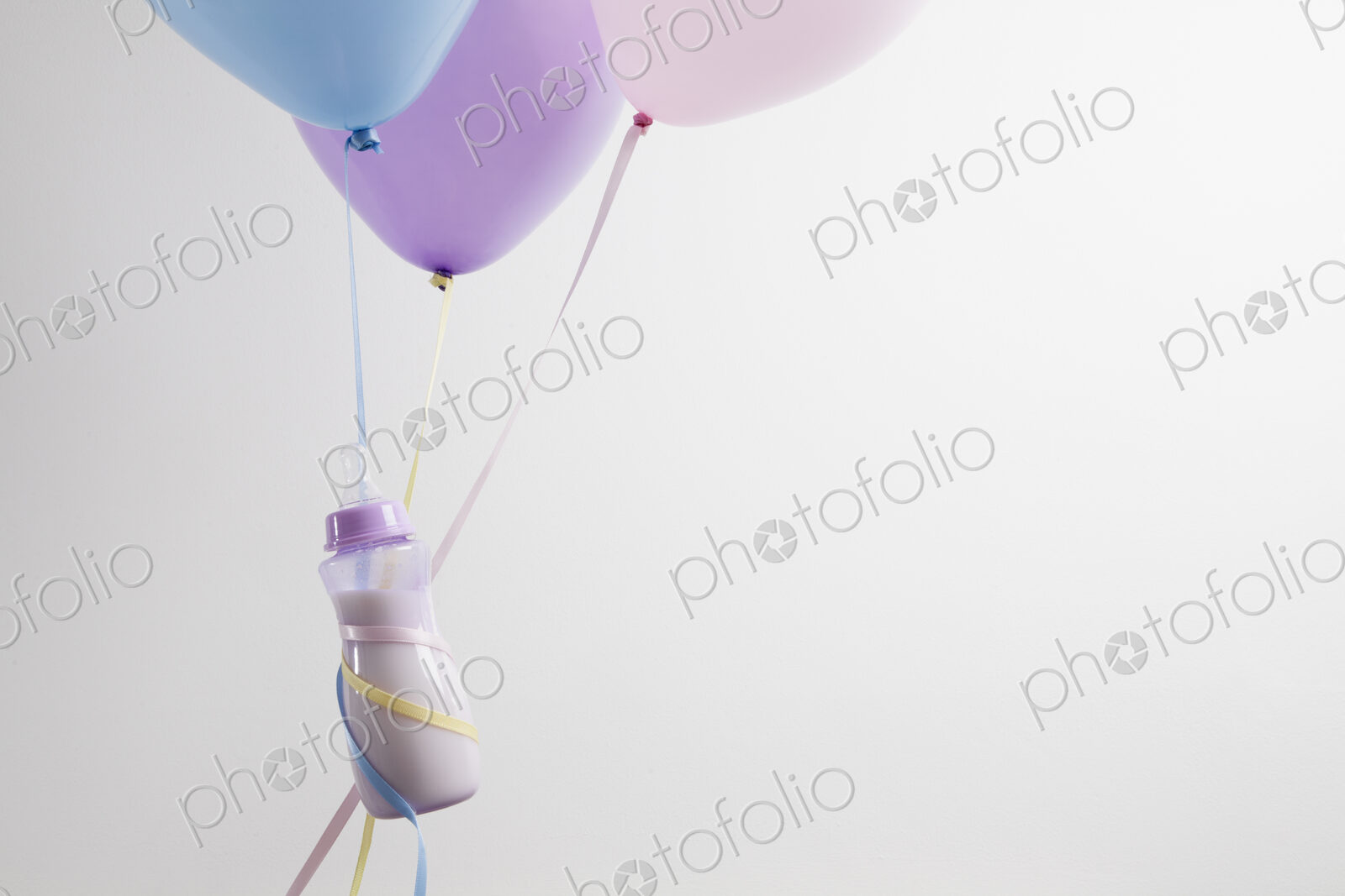A baby bottles ties with ribbon to three balloons, floating.