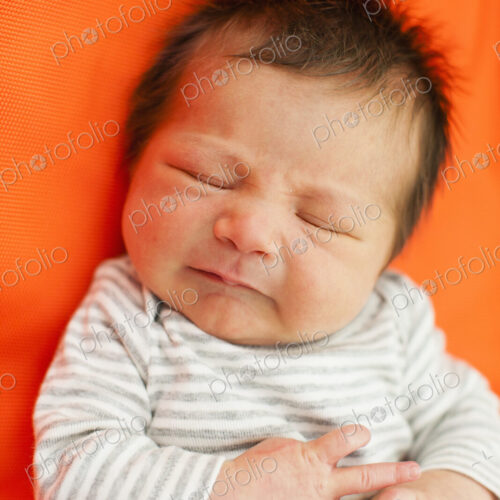 A baby in a striped top with his eyes closed, lying on an orange pillow.