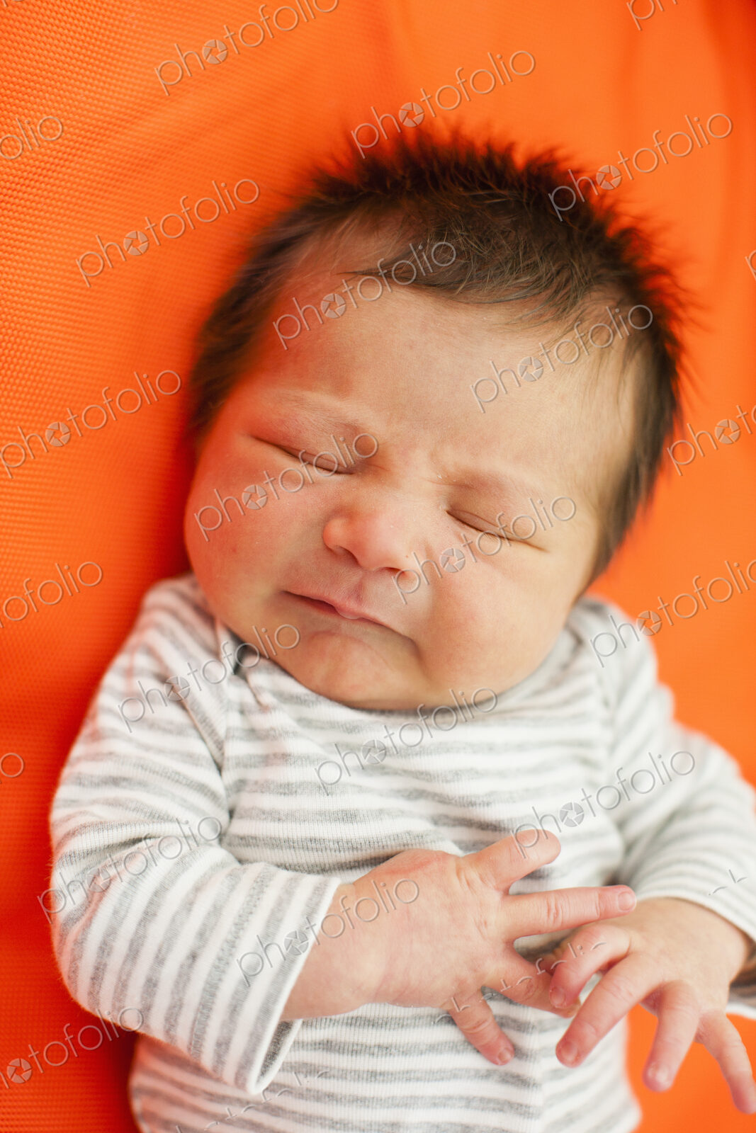 A baby in a striped top with his eyes closed, lying on an orange pillow.