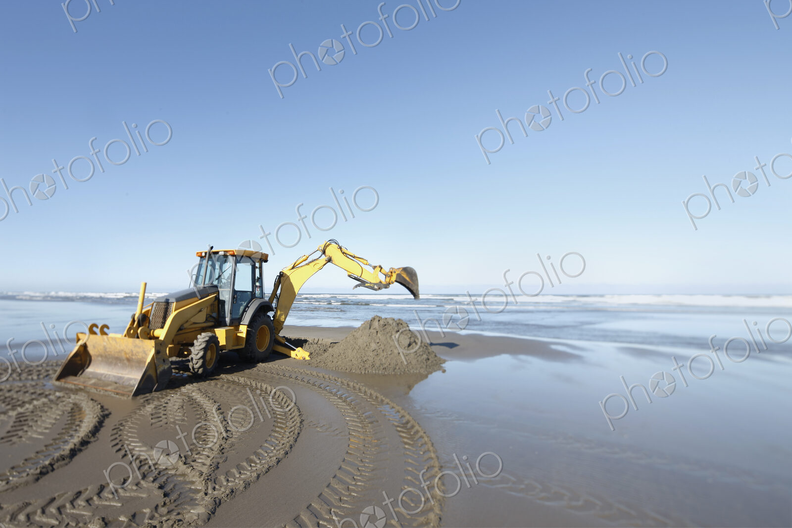 A backhoe, digger with large scoop working on the soft sand at the water’s edge.