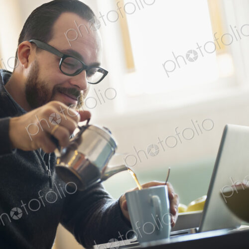 A bearded man having a drink of coffee. An open laptop computer on the counter.