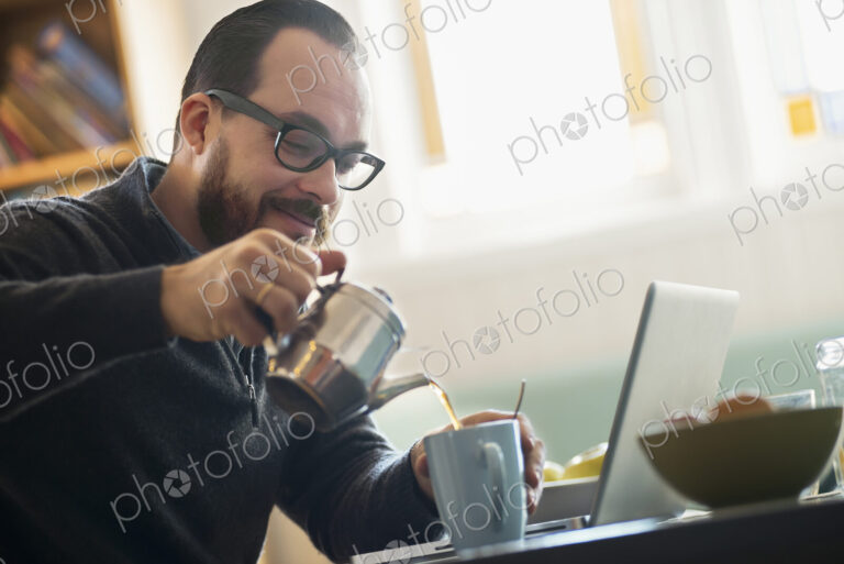 A bearded man having a drink of coffee. An open laptop computer on the counter.