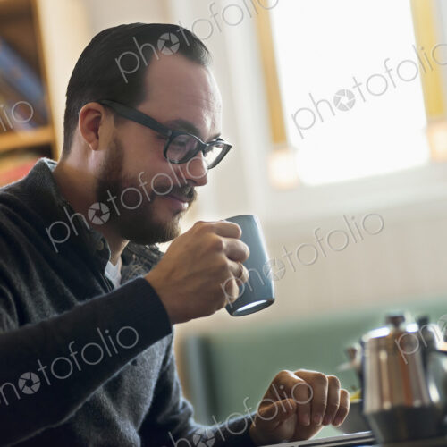 A bearded man having a drink of coffee. An open laptop computer on the counter.