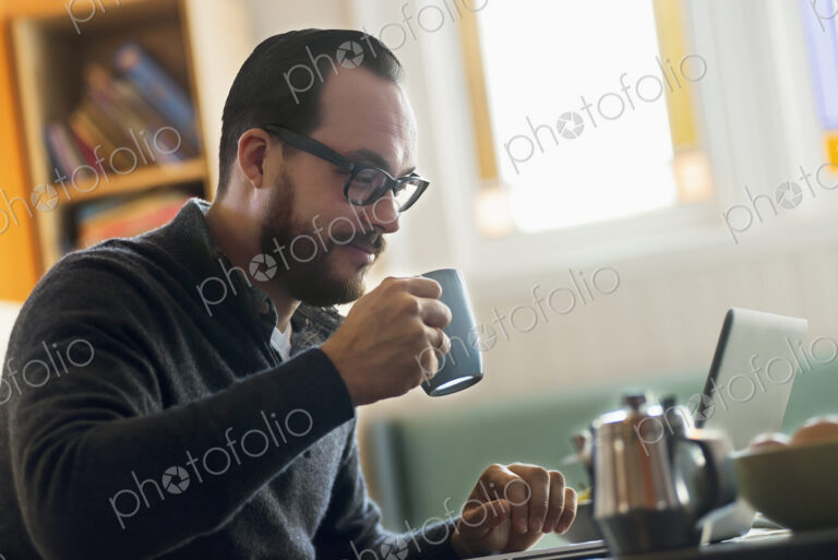 A bearded man having a drink of coffee. An open laptop computer on the counter.