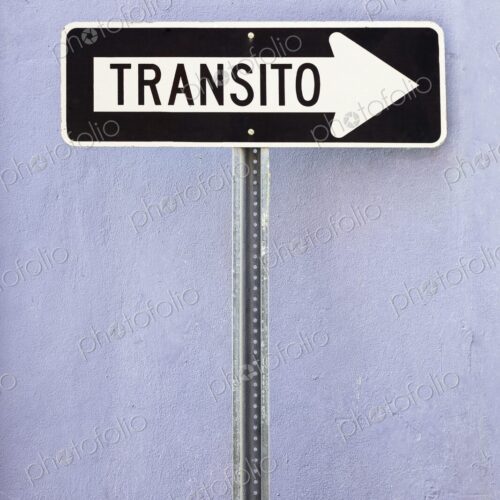 A black and white Spanish-language one-way traffic sign reading Transito is mounted… A black and white Spanish-language one-way traffic sign reading Transito is mounted…