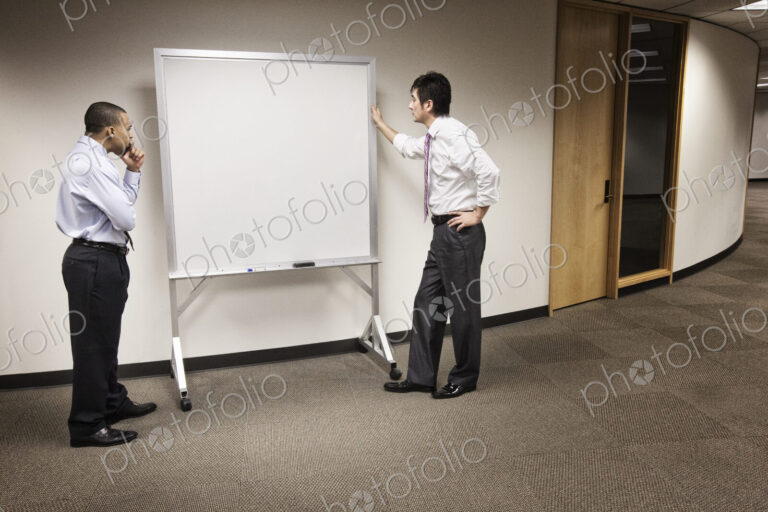 A black businessman and an asian businessman ponder the issue of an empty white board.