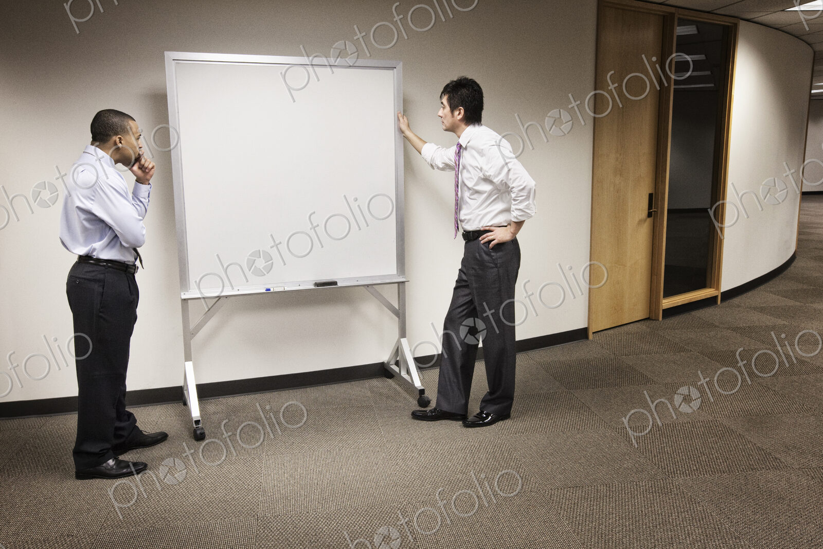 A black businessman and an asian businessman ponder the issue of an empty white board.