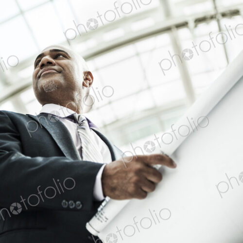 A black businessman holding plans for a new office space in a large glass covered walkway. A black businessman holding plans for a new office space in a large glass covered walkway.