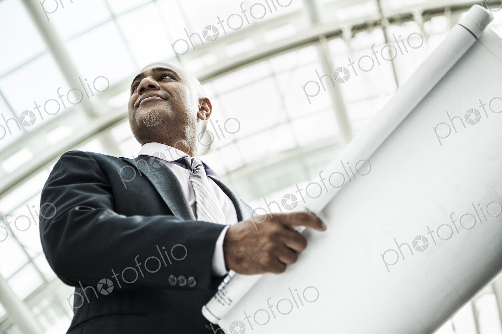 A black businessman holding plans for a new office space in a large glass covered walkway.