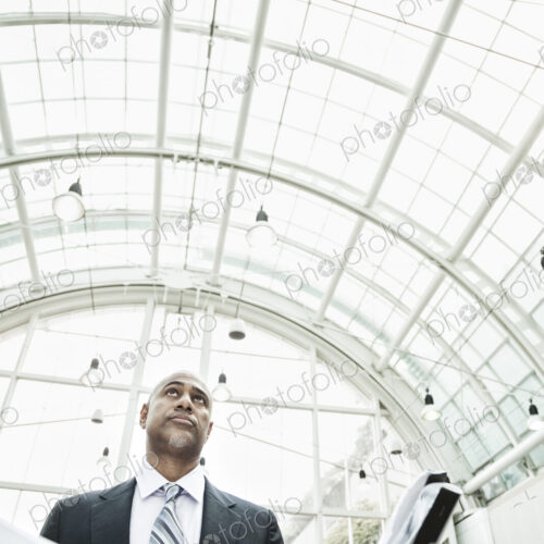 A black businessman holding plans for a new office space in a large glass covered walkway. A black businessman holding plans for a new office space in a large glass covered walkway.