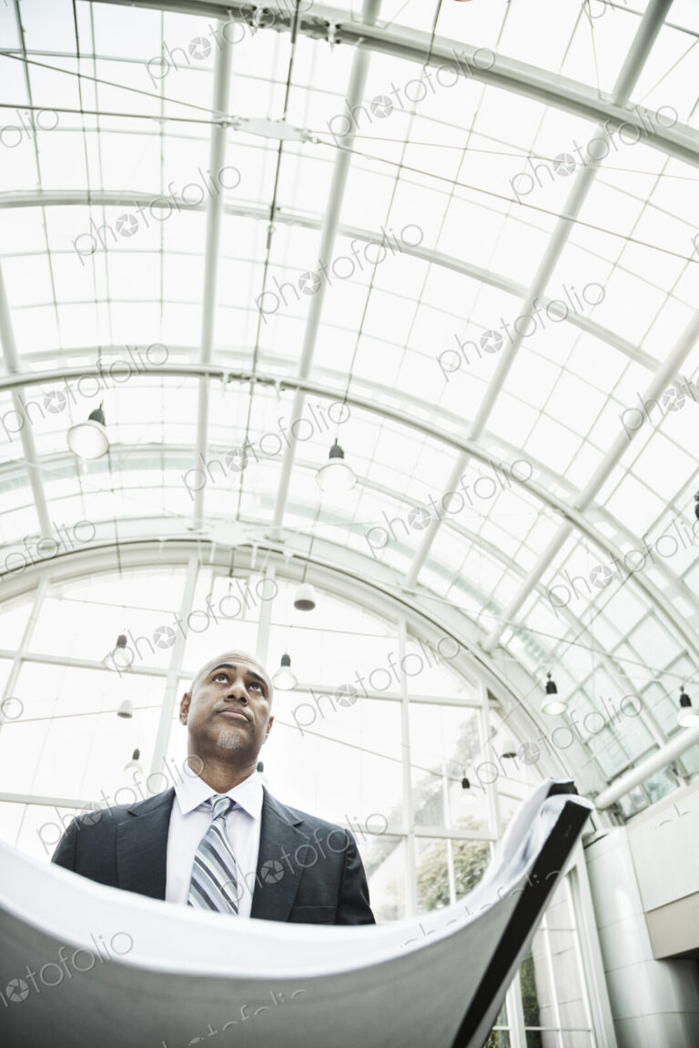 A black businessman holding plans for a new office space in a large glass covered walkway.