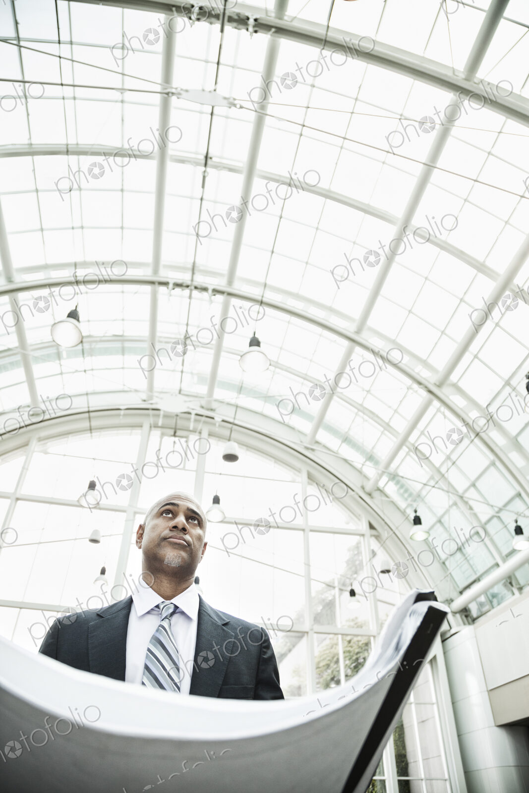A black businessman holding plans for a new office space in a large glass covered walkway.