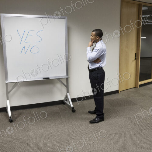 A black businessman musing by an office white board with the words Yes and No written… A black businessman musing by an office white board with the words Yes and No written…