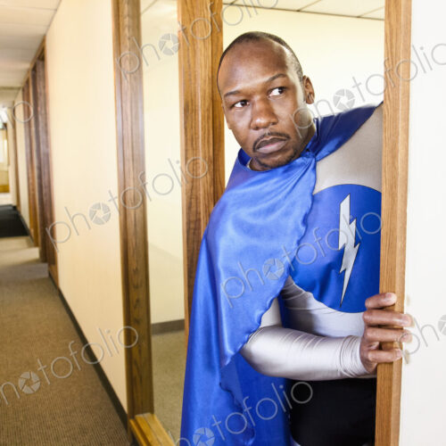 A black businessman office super hero peers out from his office into a hallway.