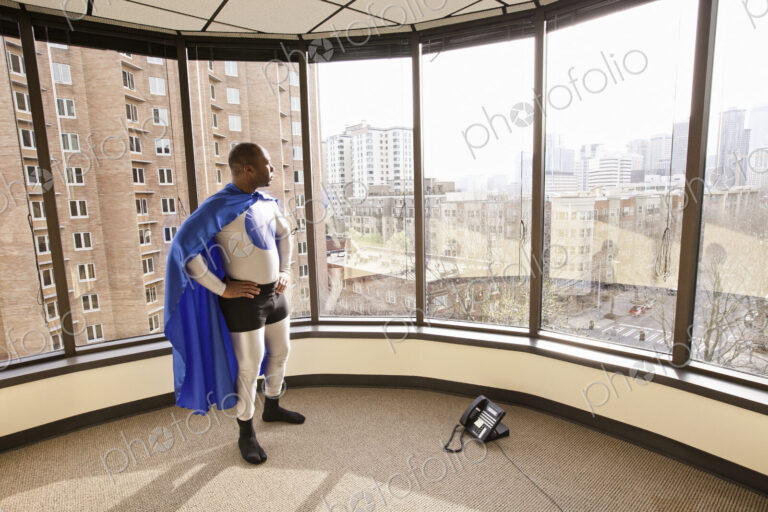 A black businessman office super hero stands at an office window and ponders his next…