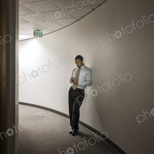 A black businessman standing alone checking his cell phone in an office hallway. A black businessman standing alone checking his cell phone in an office hallway.