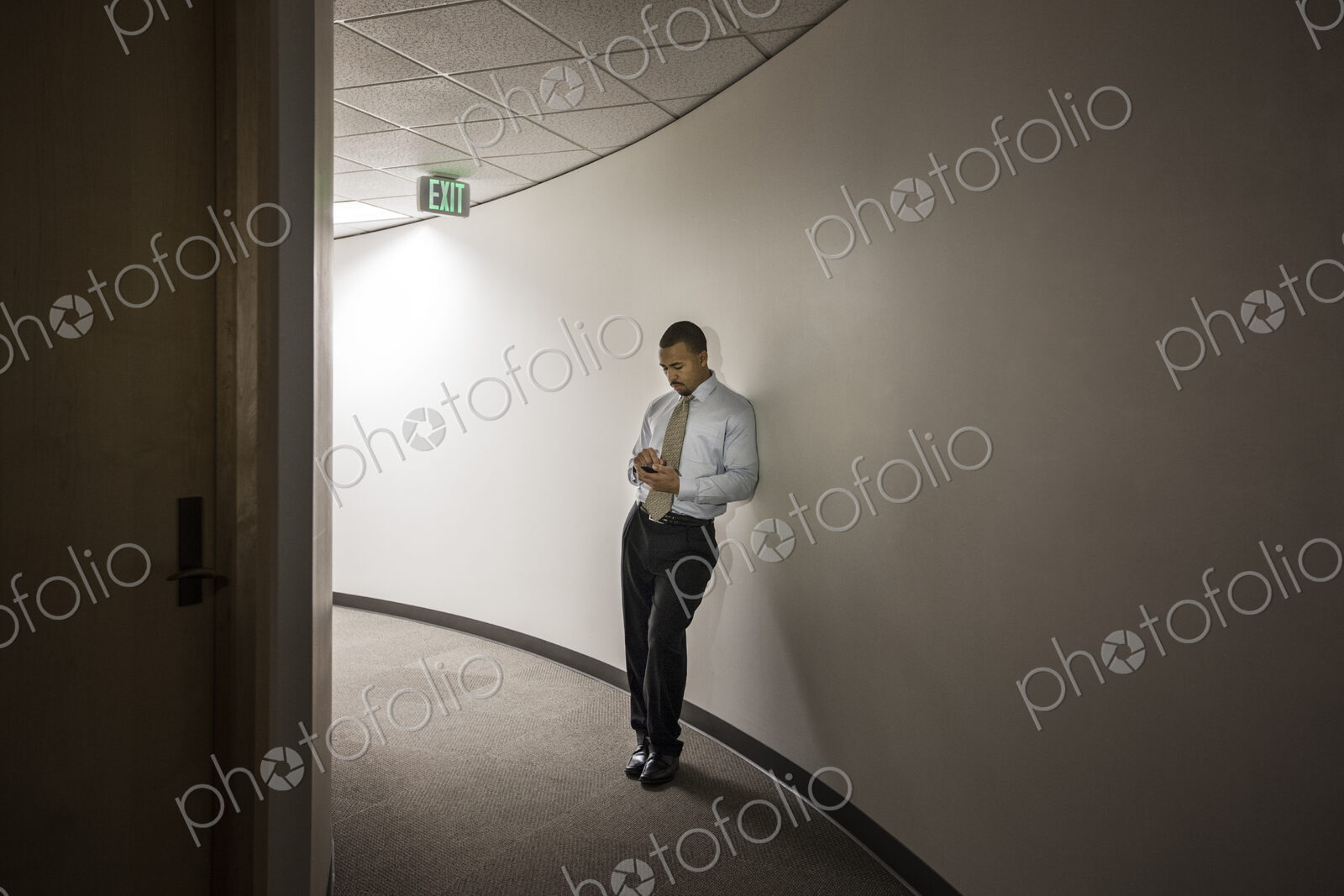 A black businessman standing alone checking his cell phone in an office hallway.