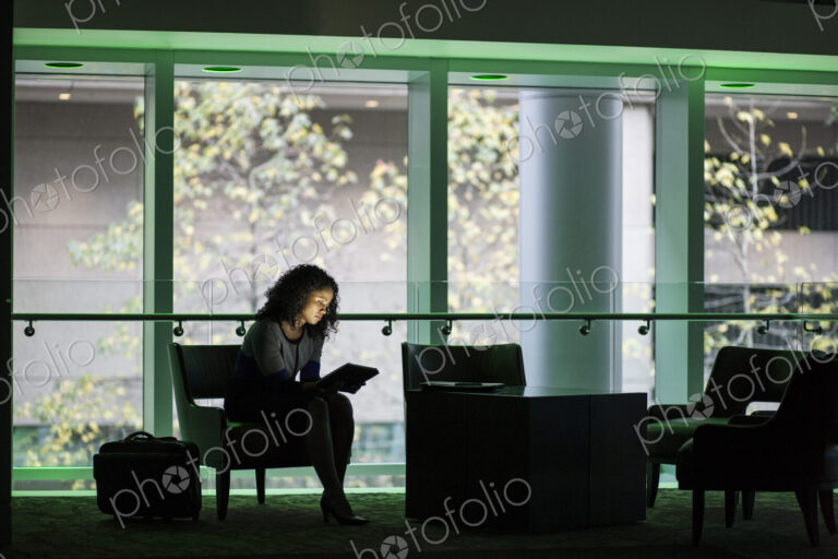 A black businesswoman sitting in front of windows in a darkened lobby area of a…