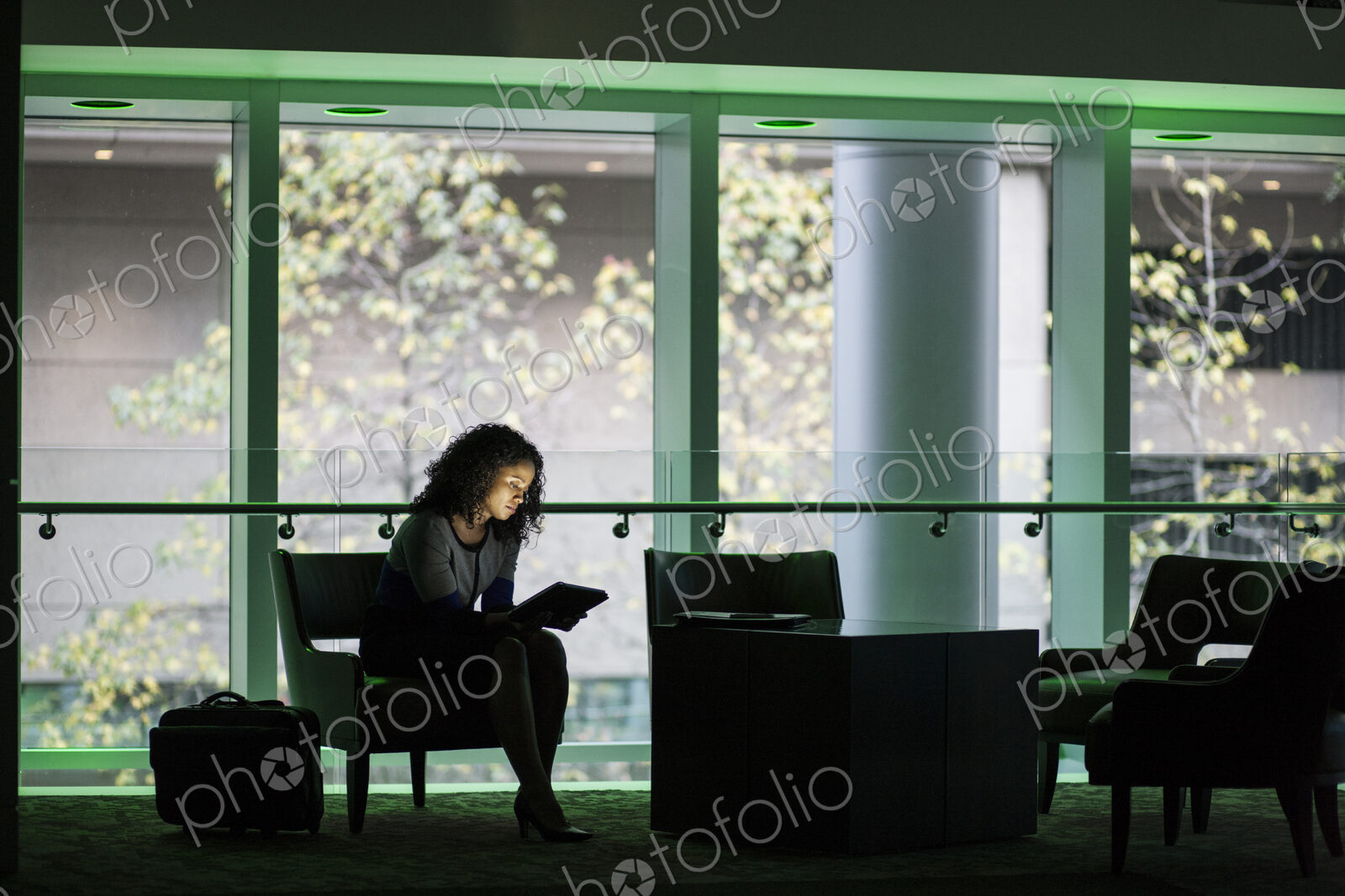 A black businesswoman sitting in front of windows in a darkened lobby area of a…