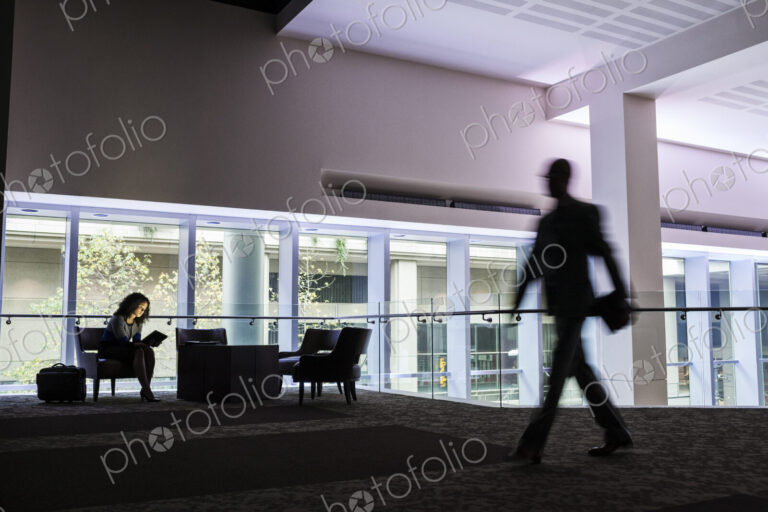 A black businesswoman sitting in front of windows in a darkened lobby area of a…