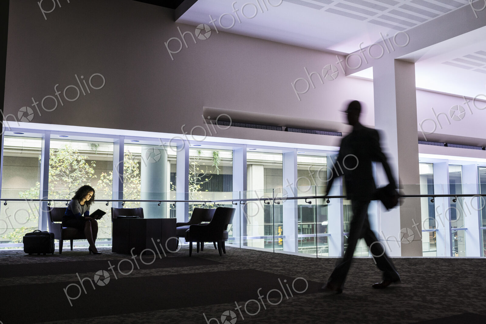 A black businesswoman sitting in front of windows in a darkened lobby area of a…
