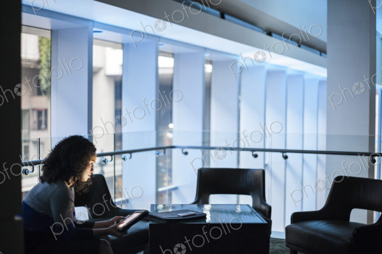 A black businesswoman sitting in front of windows in a darkened lobby area of a…